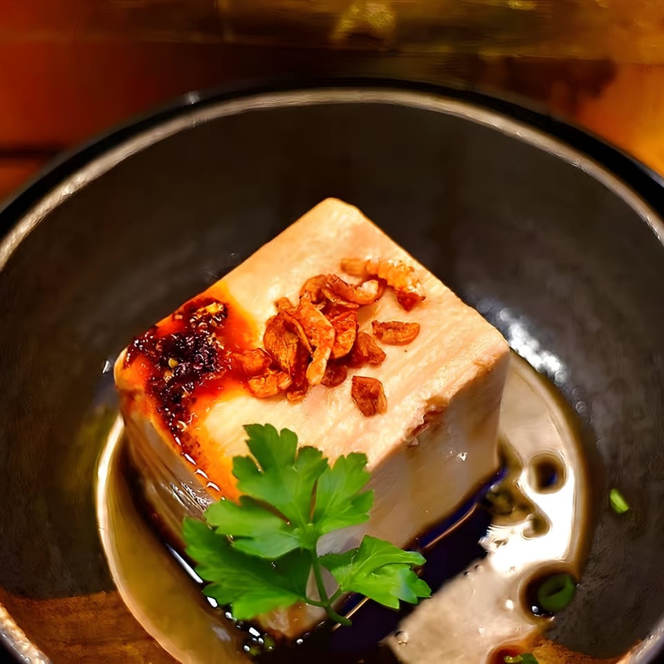 A square of BAHT Thai tofu with chili oil, crispy shallots, and parsley in a dark bowl.