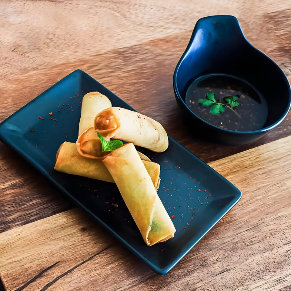 Crispy Thai spring rolls on a blue plate with a side of dark dipping sauce.