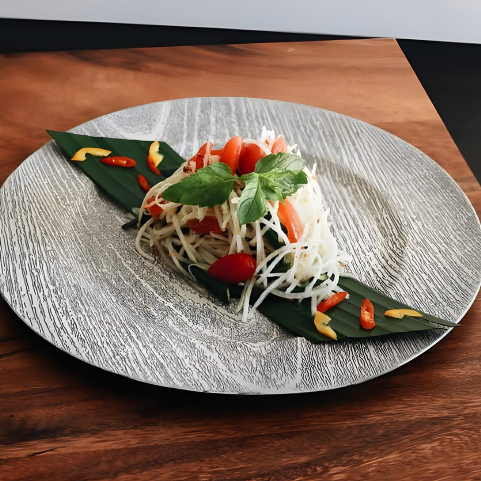 Thai papaya salad with tomatoes, basil, and chilies on a banana leaf, served on a textured plate.